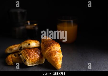 Colazione con croissant, dolci alla cannella e cioccolato, succo d'arancia fresco e caffè sul lato su uno sfondo scuro in una luce spot. Foto Stock