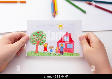 Bambino che tiene un disegno con una famiglia felice e un albero della mela e una casa. Primo piano. Foto Stock