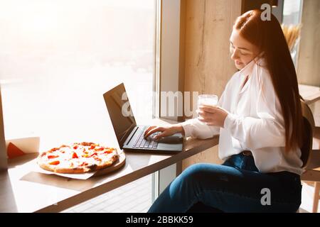 Il modello femminile funziona su un computer portatile e parla al telefono. Giovane donna beve caffè in un caffè e fare un affare. Foto Stock