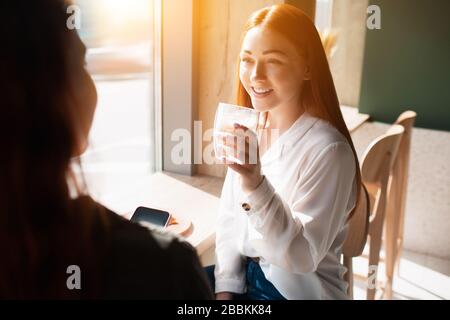 Giovane donna tiene una tazza di caffè e parla con la sua amica. Il modello femminile beve il caffè e si siede in una caffetteria Foto Stock