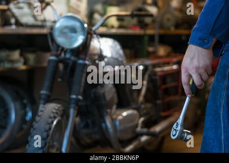 Mano dell'uomo che tiene una chiave a bussola metallica con cricchetto in garage, sullo sfondo della motocicletta Foto Stock