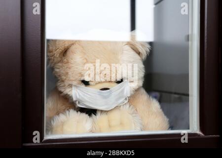 Orsacchiotto in una maschera medica viso guarda fuori dalla finestra. Concetto di quarantena del coronavirus durante l'epidemia di COVID-19 Foto Stock