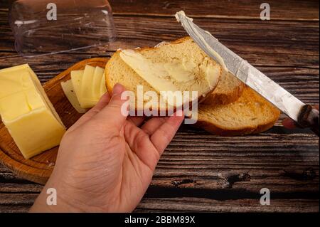 qualcuno spalma il burro su pane tostato di grano fresco con un coltello, un piatto di burro di legno con burro e fette di formaggio su uno sfondo di legno. Primo piano Foto Stock