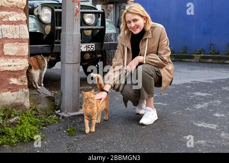 Sorridente giovane viaggiatore donna che ha strokato un gatto randagio con zenzero in pastina sulla strada di Istanbul. La femmina caucasica viaggia in Turchia. Foto Stock