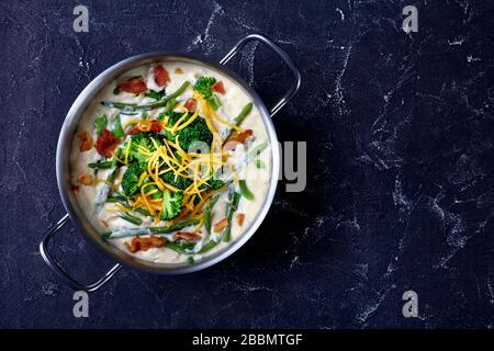 deliziosa zuppa di panna con broccoli, fagiolo verde, pancetta fritta e formaggio cheddar grattugiato in una casseruola di metallo su un tavolo di cemento, piatto, spac libero Foto Stock