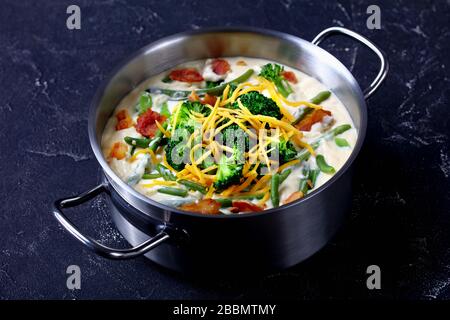 deliziosa zuppa di panna con broccoli, fagiolo verde, pancetta fritta e formaggio cheddar grattugiato in una casseruola di metallo su un tavolo di cemento, primo piano Foto Stock