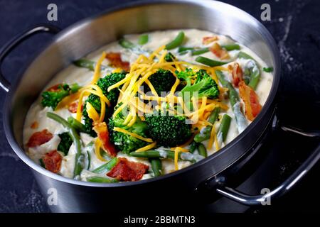 primo piano di deliziosa zuppa di panna con broccoli, fagiolo verde, pancetta fritta e formaggio cheddar grattugiato in una casseruola di metallo su un tavolo di cemento Foto Stock