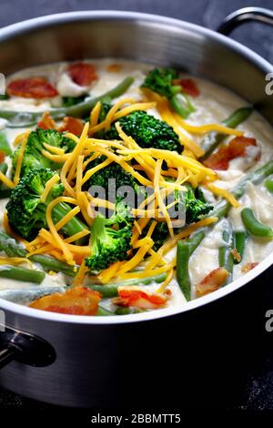 deliziosa zuppa di panna con broccoli, fagiolo verde, pancetta fritta e formaggio cheddar grattugiato in una casseruola di metallo su un tavolo di cemento, vista verticale, clos Foto Stock