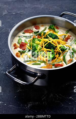 deliziosa zuppa di panna con broccoli, fagiolo verde, pancetta fritta e formaggio cheddar grattugiato in una casseruola di metallo su un tavolo di cemento, vista verticale Foto Stock