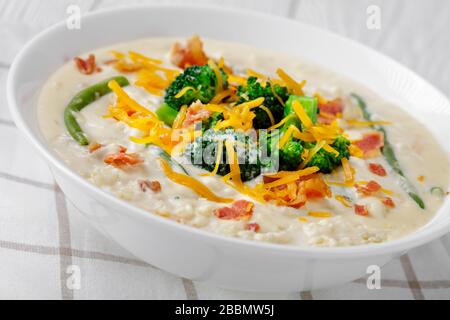 primo piano di una porzione di zuppa di panna con broccoli, fagiolo verde, pancetta fritta e formaggio cheddar grattugiato in una ciotola bianca su un tavolo di legno, orizzontale vi Foto Stock