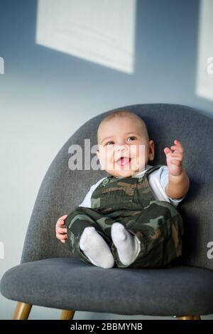 ragazzino di 8 mesi di età in tute, una camicia bianca e calze bianche sedute su una sedia grigia Foto Stock