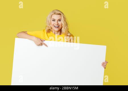 Giovane donna felice si porca fuori da dietro un banner bianco su uno sfondo giallo. Selezionare uno spazio vuoto su un modulo, uno spazio di copia per il testo. Orizzontale sh Foto Stock