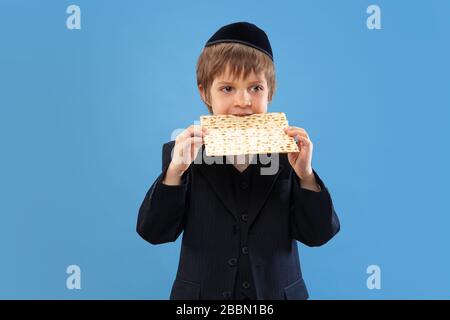 Joyfull mangiare matzah. Ritratto di un giovane ebreo ortodosso isolato su sfondo blu studio. Purim, business, festival, vacanza, infanzia, celebrazione Pesach o Pasqua, giudaismo, religione concetto. Foto Stock