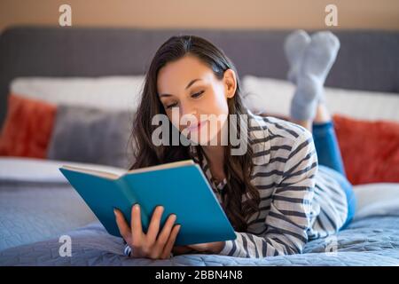 Giovane donna felice sdraiata a letto nella sua casa e libro di lettura. Foto Stock