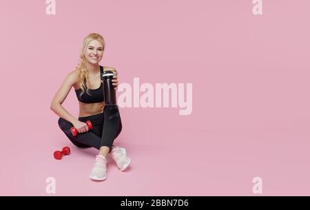 Training personale pubblicitario. Una giovane donna sportiva beve un frullato proteico da un agitatore dopo l'allenamento al chiuso. Sfondo rosa Foto Stock