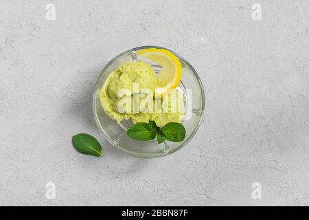 Dolce vegano fatto in casa gelato avocado con foglie di limone e menta in una ciotola di vetro su sfondo grigio. Vista dall'alto con spazio di copia Foto Stock