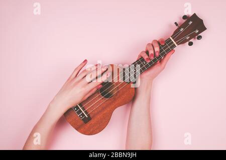 Donna mani con bellezza rosa manicure tenendo piccolo wodden ukulele su sfondo rosa. Bella foto di musicista femminile. Foto Stock