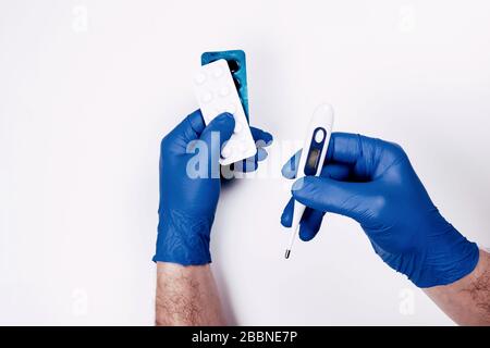 Le mani dell'uomo in guanti di smaltimento blu tenere termometro e pillole di medicina su sfondo bianco. Trattamento medico di emergenza. Foto Stock