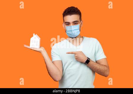 Ritratto di giovane bruna felice con maschera medica chirurgica che punta alla casa di carta sul palmo e sorridente alla macchina fotografica, acquisto domestico. ind Foto Stock
