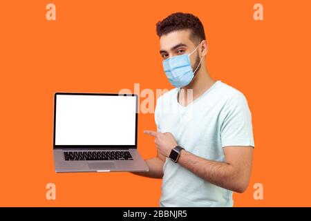 Ritratto di un uomo bruna gioioso positivo con maschera chirurgica medica che tiene in piedi il laptop con schermo vuoto e sorridente alla macchina fotografica, advertis del Internet Foto Stock