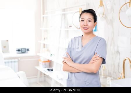 Ritratto di estetista asiatico positivo di mezza età in scrub in piedi con braccia incrociate in sala trattamento di bellezza con design contemporaneo Foto Stock