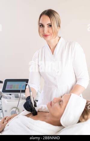 Ritratto di contenuto sicuro specialista di bellezza in camice da laboratorio utilizzando la macchina cosmetologica per lisciare la pelle sul petto dei clienti Foto Stock