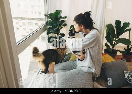 Ragazza hipster abbracciando e giocando con due gatti nella stanza moderna, seduta insieme a casa durante la quarantena di coronavirus. Soggiorno a casa sicuro. Isolamento Foto Stock