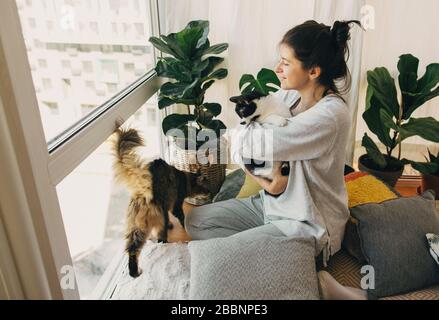 Soggiorno a casa sicuro. Ragazza hipster abbracciando e giocando con due gatti nella stanza moderna, seduta insieme a casa durante la quarantena di coronavirus. Isolamento Foto Stock