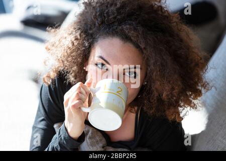 Giovane e bella donna bruna con capelli ricci si trova sul divano, sorseggiando dalla tazza bianca e guardando la macchina fotografica. Primo piano verticale Foto Stock