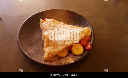 Frittella / Crepe primo piano con frutta e zucchero di pasticceria come banana , fragola su un piatto su un tavolo scuro - dolce fatto in casa o colazione Foto Stock