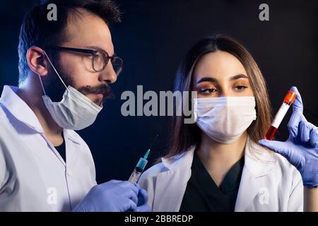 La donna bianca caucasica del medico o dell'infermiere contiene una provetta per il campione di sangue e un medico maschio con occhiali per gli occhi contiene siringa per iniezione e vaccino. Foto Stock