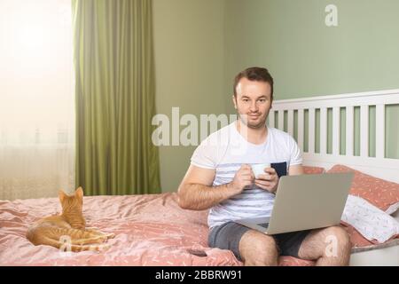 Giovane con un computer portatile a letto che lavora a casa con una tazza di caffè. Un allegro adulto che lavora dal suo letto di appartamento con un gatto accanto a lui sul letto. Foto Stock