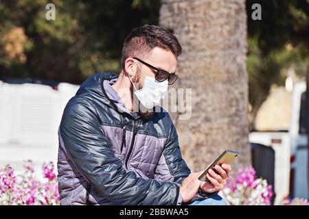 La maschera da uomo usa lo smartphone seduto nel parco all'aperto. Autoprecauzioni durante la pandemia di Coronavirus. Foto Stock