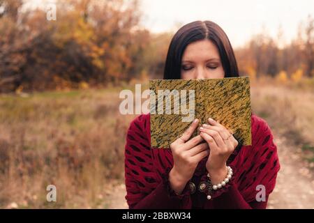 Una ragazza in un abito retrò tiene un libro aperto che copriva metà della sua faccia. C'è molto spazio per il design sulla metà sinistra del telaio. Nostalgia autunno umore. Foto Stock