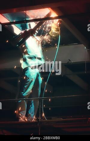 Saldatrice professionale in uniforme protettiva e maschera per saldatura di metalli nell'oggetto industriale. Foto Stock