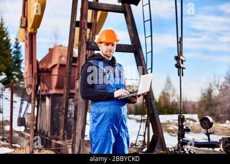 Ritratto di ingegnere di petrolium, indossando tute blu e casco arancione, con un laptop, in piedi con la schiena a un carro di perforazione, controllando l'unità di pompaggio dell'olio, prendendo appunti nel suo computer Foto Stock