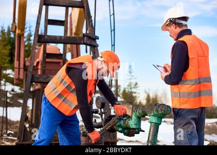 Due operai dell'olio che indossano le tute, i giubbotti arancioni ed i caschi, che lavorano sul campo dell'olio vicino al martinetto della pompa dell'olio, uno sta strappando il tubo, l'altro fa le note. Concetto di industria petrolifera, collaborazione Foto Stock