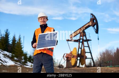 Uomo d'affari sorridente in gilet arancione e casco bianco che tiene mini pannello solare in calda giornata di sole, sullo sfondo il jack pompa e cielo blu, concetto di fonte alternativa di energia Foto Stock