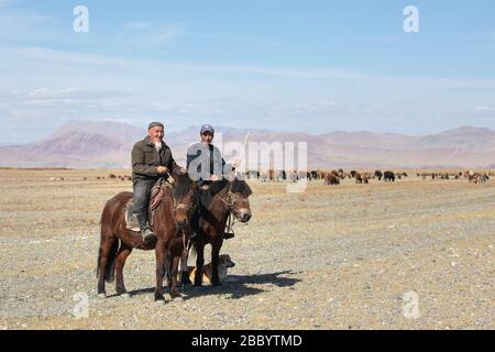 Ulgii, Mongolia - 21 settembre 2019: Due pastori mongoli a cavallo che erigono pecore con il loro cane. Foto Stock