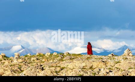 Donna, pellegrino buddista in veste rossa camminando nel deserto. Sullo sfondo montagne dell'altopiano tibetano. Vicino al lago Nam Co. Cielo drammatico Foto Stock