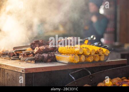 Street food sulla griglia. Spiedini di carne e mais, barbecue. Sullo sfondo sono le mani di un uomo con un coltello che sta cucinando kebab. Esterno. Foto orizzontale. Foto Stock