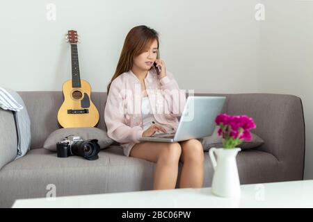 Giovane donna freelance asiatica che parla con lo smartphone e utilizza il computer portatile mentre si siede sul divano a casa. Foto Stock