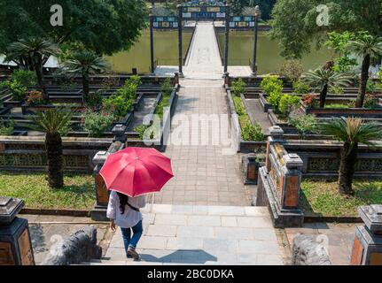 Signora vietnamita con ombrellone che cammina verso il ponte Trung Dao e la tomba di Minh Mang nei giardini del complesso della cittadella imperiale Hue , Vietnam Foto Stock