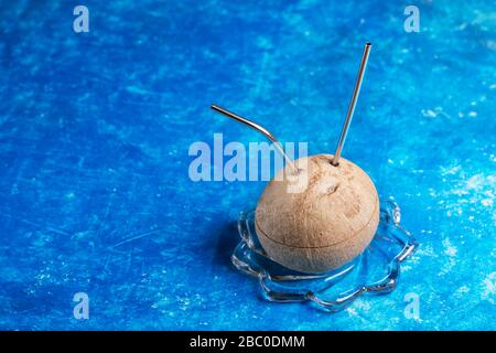 Cocco fresco con due cannucce metalliche inserite in esso su lastra di vetro su sfondo blu. Zero rifiuti e nessun concetto di plastica. Rifiuto del bere di plastica Foto Stock