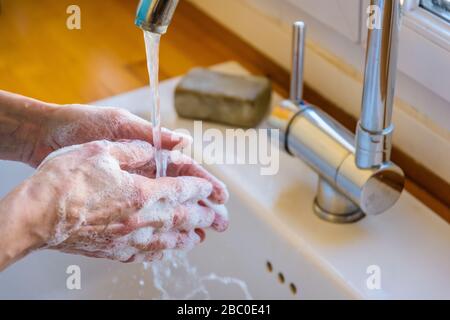 Vista ravvicinata sulle mani di una donna lavando accuratamente le mani con sapone sotto il rubinetto del lavello della cucina. Foto Stock