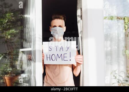 Un giovane uomo in una maschera protettiva si alza a casa fuori dalla finestra e mostra un tavolo con le parole rimanere a casa. Una richiesta di autoisolamento a causa di una pandemia e del rischio di infezione da coronavirus. Foto Stock