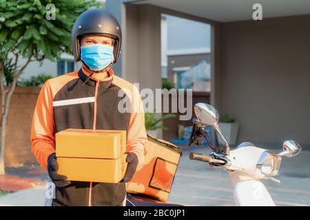 Asian consegna giovane uomo con maschera protettiva in uniforme arancione reggendo pile di scatole di cartone in casa villaggio di fronte con scooter. Pubblicità, Busi Foto Stock