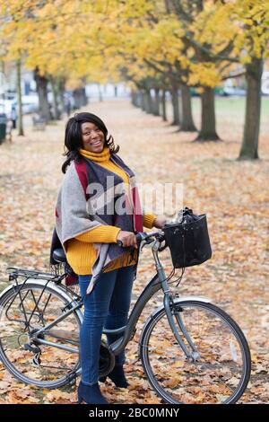 Ritratto sorridente, sicura donna in bicicletta tra gli alberi e le foglie nel parco autunnale Foto Stock