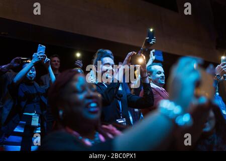 Un pubblico sorridente ed entusiasta che utilizza telefoni con fotocamera nell'auditorium scuro Foto Stock