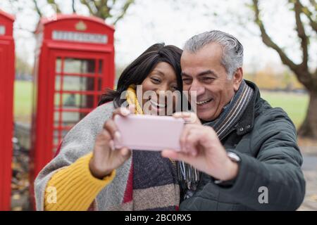 Coppia senior sorridente che prende il selfie nel parcheggio di fronte alla cabina telefonica rossa Foto Stock